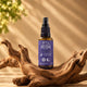 Flacon spray en verre ambré 30 ml de Reishi Pure Croft avec embout pulvérisateur noir, posé sur du bois flotté naturel, formule routine nocturne au zeste d'orange.