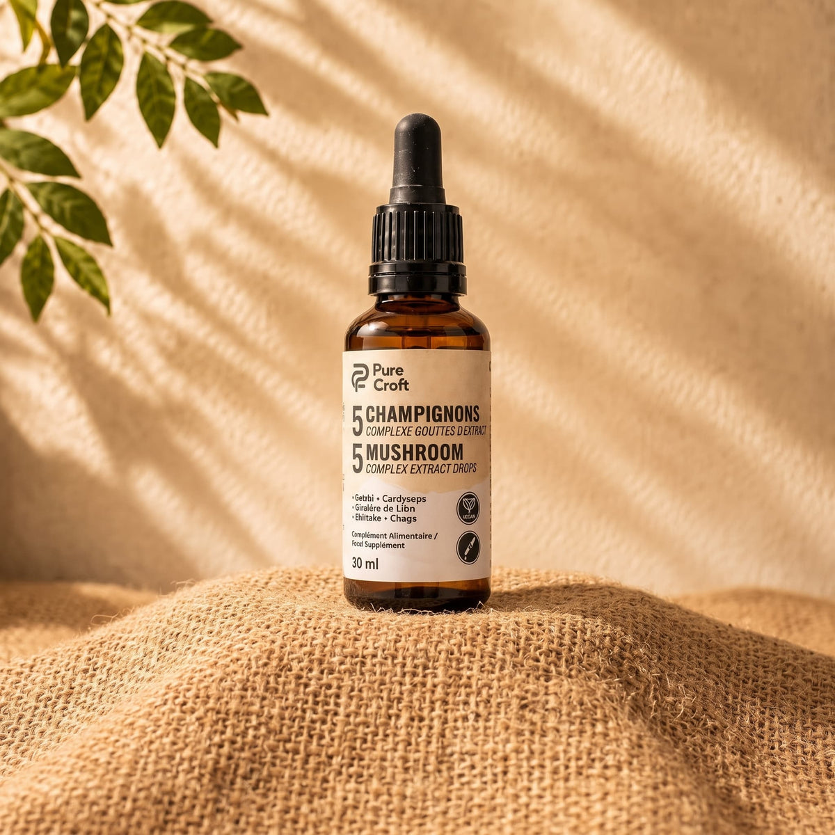 Un flacon unique de 30ml de Pure Croft Gouttes d'Extrait Complexe 5 Champignons avec une pipette noire est posé sur un tissu en toile de jute texturé. L'arrière-plan présente une lumière naturelle et des feuilles vertes.